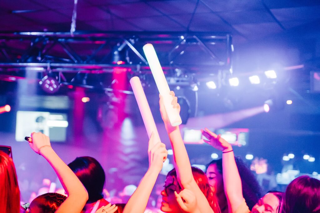 Hands holding glow sticks in the air under pink and purple lights at a Senior Week glow party